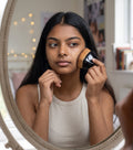Mujer joven aplicándose base con la brocha angular zafiropulse PERENNE Facet Brush frente a un espejo ovalado