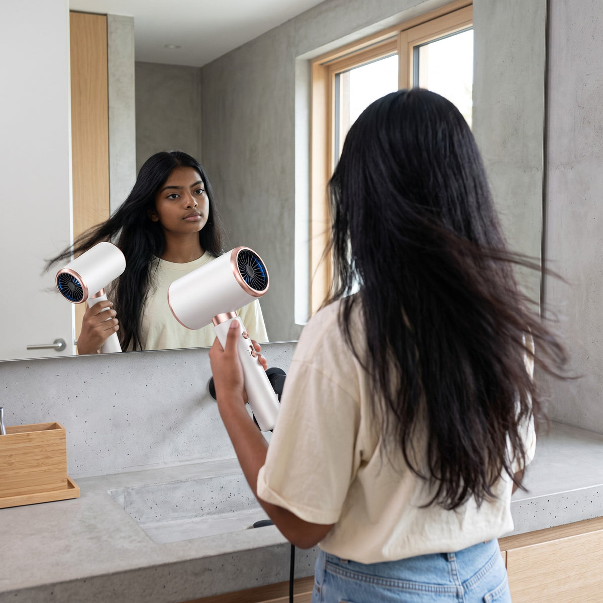 Mujer joven usando el secador de pelo zafiropulse PERENNE Aero-Glow Therapy blanco con detalles en oro rosa frente al espejo